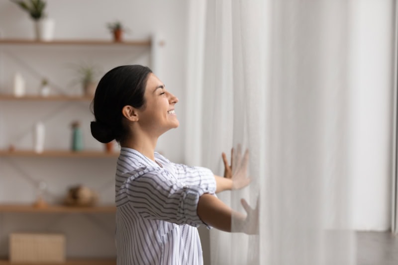 Bathing in sunlight. Excited young indian lady meet first morning at new flat house part curtains enjoy being homeowner. Happy hindu female open drapes on window breath fresh air close eyes in delight.